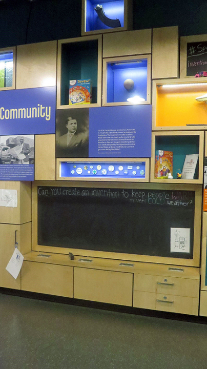 A wall of light wood cabinets and cubbies have purple or orange interiors and objects in lighted display cases. Below the cubbies and cabinets is a long rectangular blackboard and a wooden shelf underneath with more cabinets and drawers.