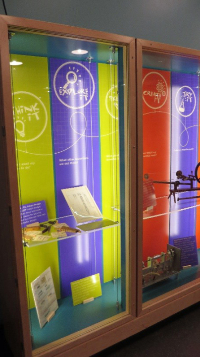 A light wooden display case with a glass cover and glass shelves holds objects, books, and papers. At the back of the display case are colorful  panels with logos that say think it, explore it, create it, and try it.