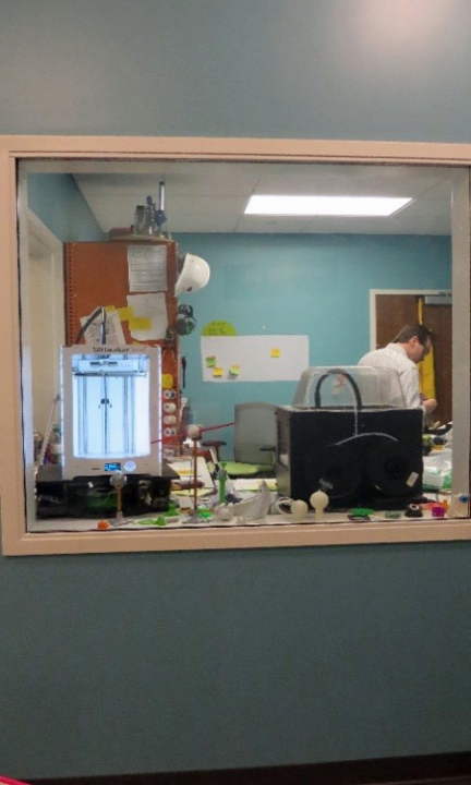 A blue wall with a clear window in the middle. Looking through the window you can see Inventor Tim and his workshop filled with tools, 3D printers on a workbench, an orange cabinet, and pieces of paper hanging on the walls.