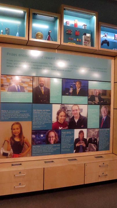 A wall in sparklab has blue text panels arranged across it. Each of the blue panels features a picture of an inventor with text about their invention.