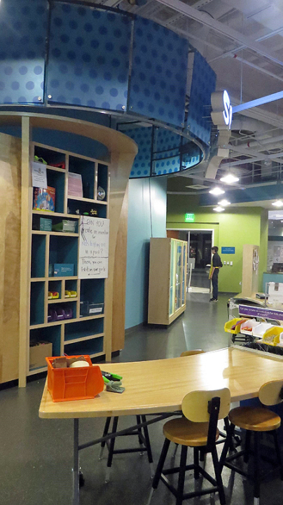 Sparklab interior taken from the back of the room. In the foreground a light wooden curved table with stools and some craft supplies in an orange bucket. Behind the table is are light wooden shelves with buckets of materials.