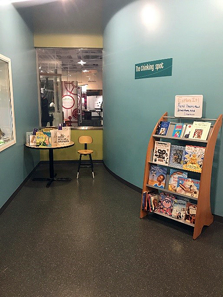 On the wall, a sign says Thinking Spot. Under the sign is a wooden book shelf with four shelves displaying children's books. In the background is a round table with more children's books on display. 