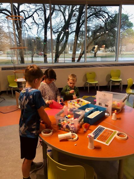 Three young inventors work on flying object prototypes together around an orange table.