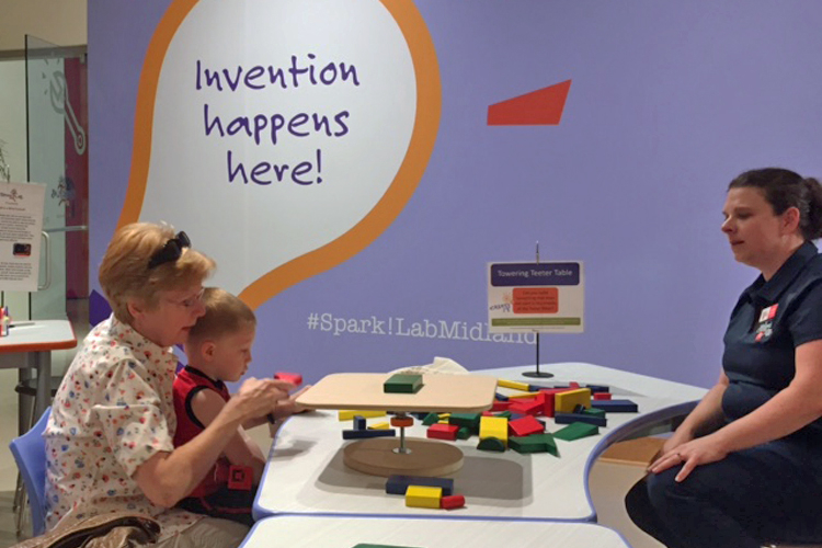 A woman holding a small child sits at a table across from a SparkLab facilitator and works on an activity at the Midland Center for the Arts