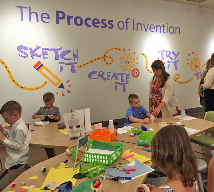 3 young boys and 1 young girl work at activity tables, using craft materials in front of them. An adult woman assists 1 boy. “The Process of Invention” is written in large letters on the wall, along with the illustrated words Sketch It! Create It! Try It!