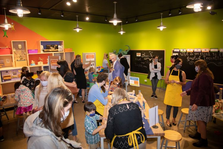 A large group of people gather in a room painted bright green and orange. There are chalkboards on the right-side walls and shelves full of books and objects on the left.