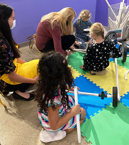 3 young girls, an adult woman, and a woman facilitator work on building a vehicle with PVC pipe