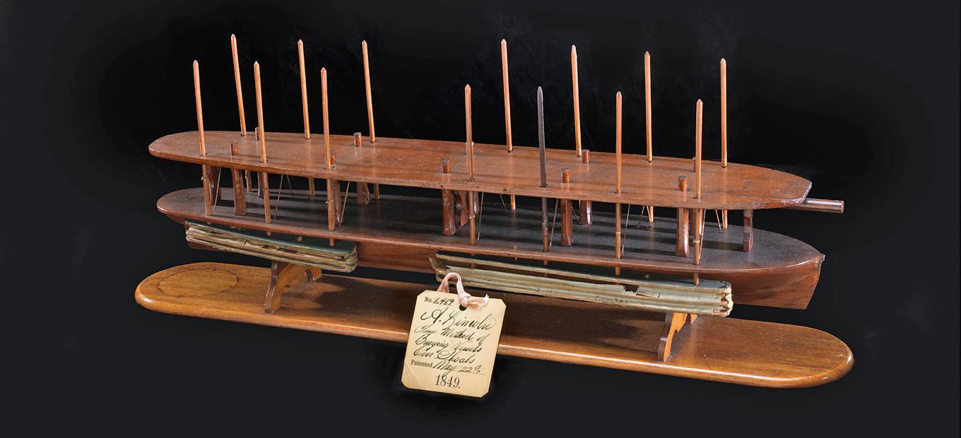 Wooden model of a long, narrow boat hull with air-filled chambers on the outside. The chambers are moved up or down with vertical poles that connect through the hull. Pushing the chambers into the water raises the bottom of the boat.