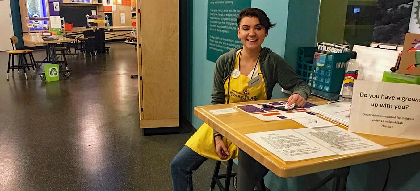 Hannah Correlli, a young woman with short, dark hair and wearing jeans, a hoodie, and a yellow SparkLab apron, sits at the welcome desk at the entrance to SparkLab, smiling into the camera.