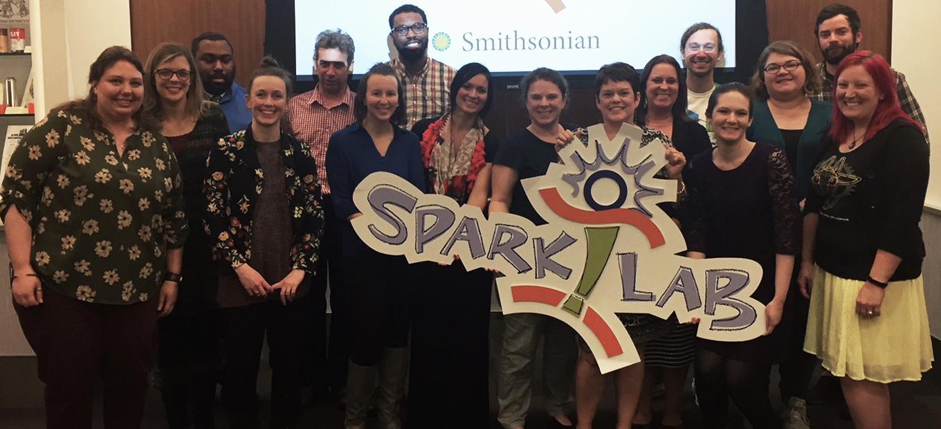 Group photo of 16 participants in the SparkLab National Network 2017 conference. Tricia Edwards holds a large cut-out of Sparky, the SparkLab logo