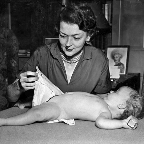 A baby holding a block in one hand lies on a table and looks up at Marion O’Brien Donovan, who is diapering the baby, using her waterproof, reusable diaper cover. She is looking down at the snaps in the cover that allow it to be adjustable to the baby’s size without the use of safety pins. She is smiling slightly and is wearing a V-neck dress and a triple strand of pearls. A framed photo of an older woman wearing a large hat is visible on a table over Donovan’s left shoulder.