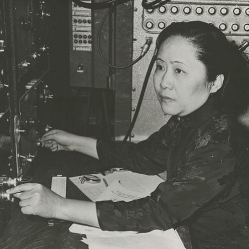 Chien-Shiung Wu in profile, sitting at a desk with a panel of knobs and dials in front of her.