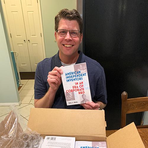 A smiling Eric Hintz holds up his book for the camera, with a full box of books in front of him