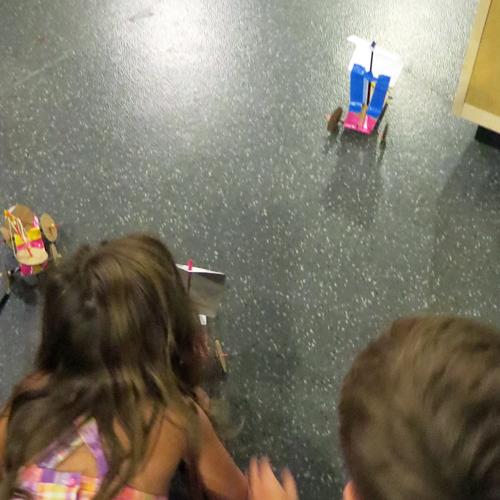 A young girl and boy are sitting on the floor, testing their inventions of vehicles with wheels and sails. The little girl seems to be blowing into the paper sail of one vehicle.